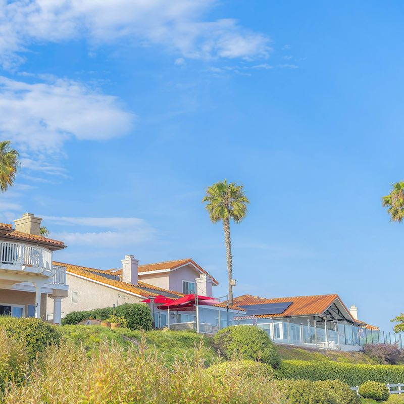 Square White puffy clouds Large residential houses and palm trees at Southern California. There is a house with terrace on the left side beside the house with solar panels and glass fence on the right.